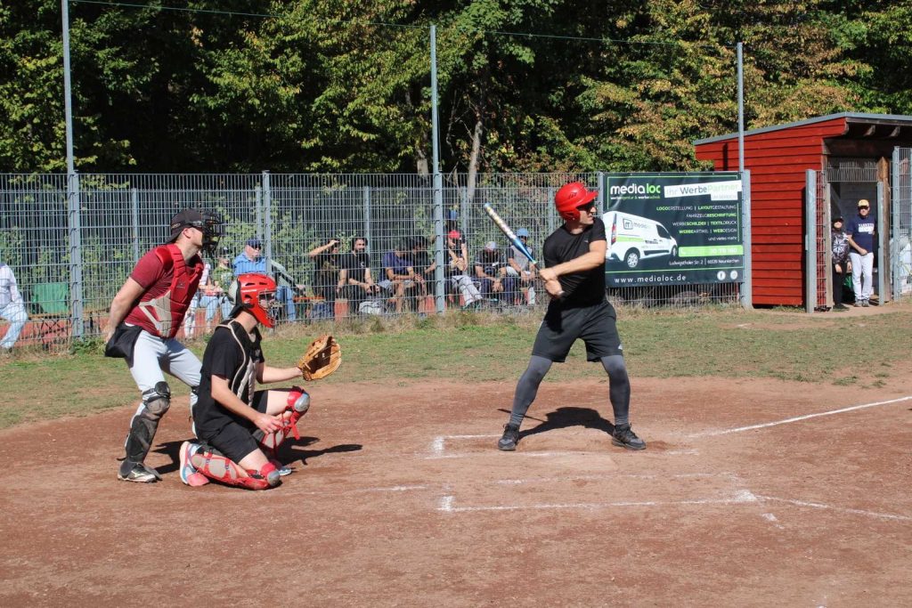 Batter mit rotem Helm wartet auf den Wurf, dahinter Catcher und Umpire