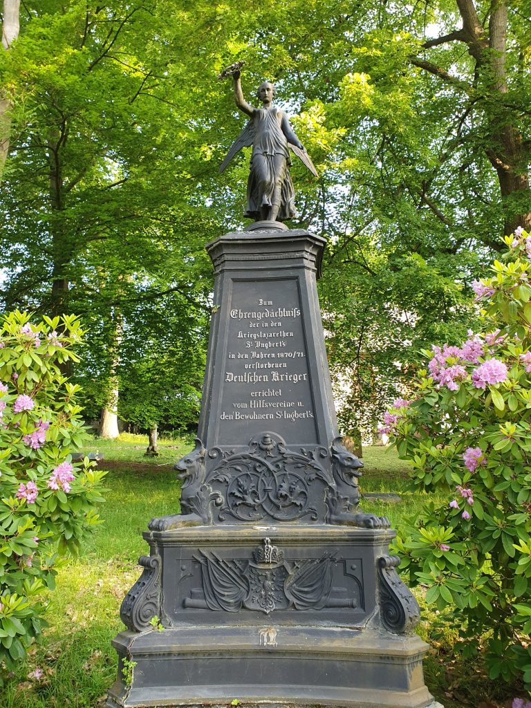 Denkmal mit Engelsfigur auf einem Sockel zwischen Bäumen und blühenden Rhododendren