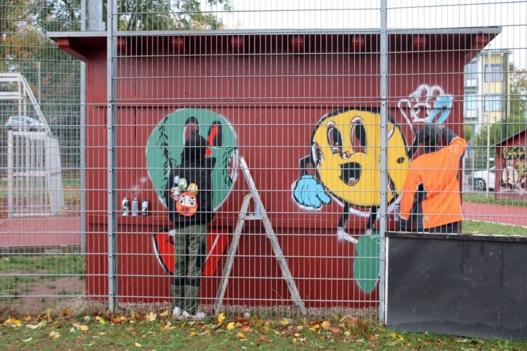 Graffiti-Figuren in Baseball-Form an einem roten Haus hinter einem Zaun