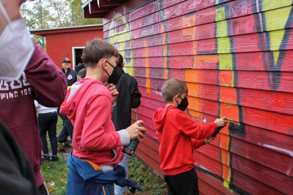 Kinder sprayen bunte Graffiti an ein Holzhaus auf dem Ballpark Wallerfeld