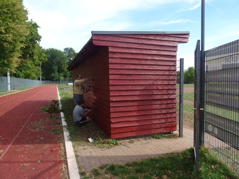 Zwei Personen streichen ein rotes Holzhaus am Rand eines Sportplatzes