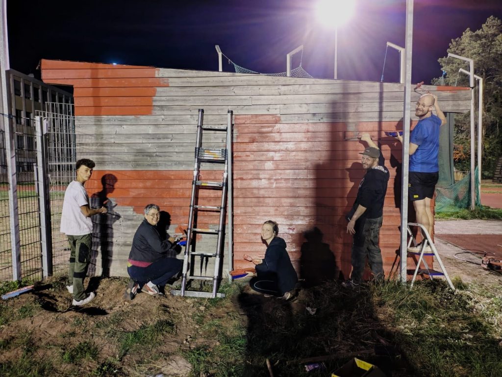 Gruppe von Vereinsmitgliedern streicht abends eine Holzwand mit roter Farbe