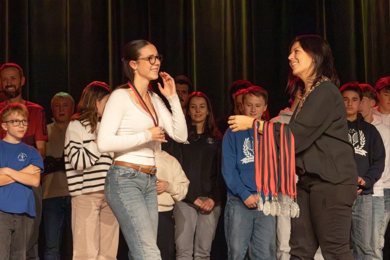 Bürgermeisterin hängt einer jungen Sportlerin auf der Bühne eine Medaille um, im Hintergrund weitere Geehrte