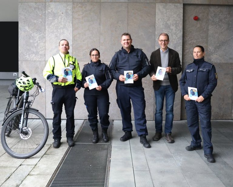 Fünf Personen, darunter Polizeibeamte und Ortspolizei, halten vor einem Gebäude den Flyer „Einbruch – nicht mit uns!“