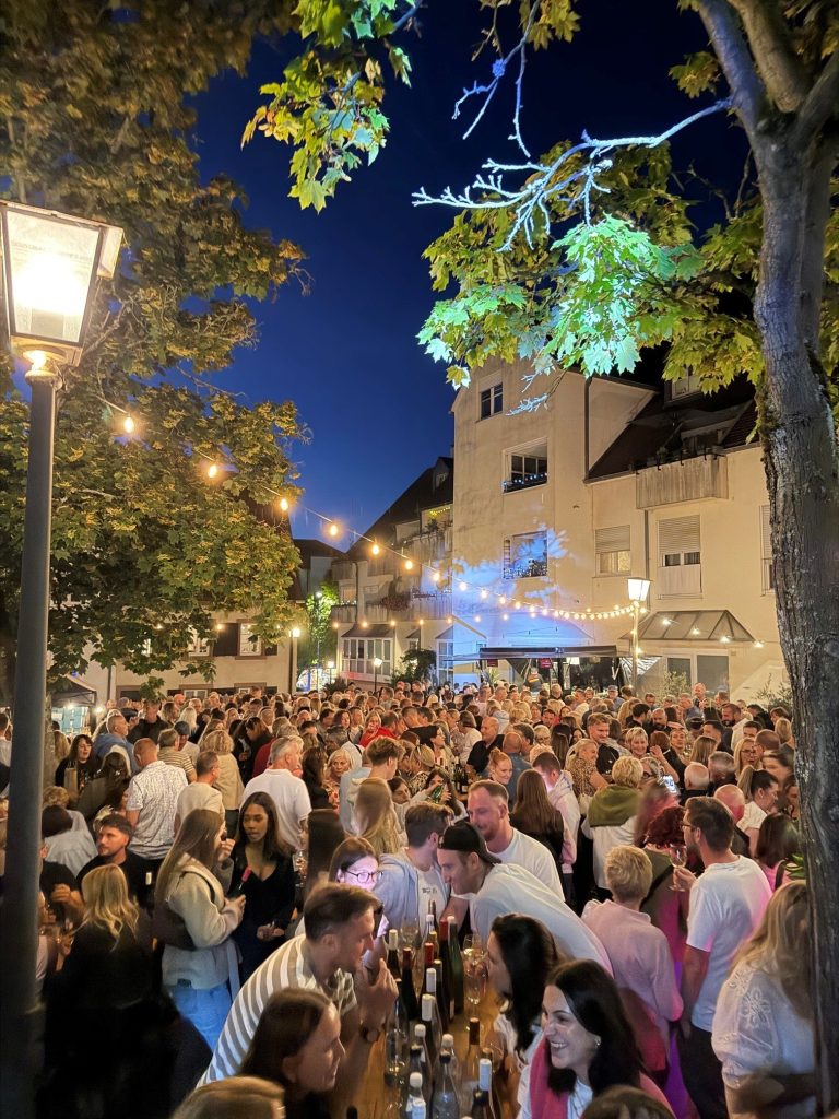 Dicht gedrängte Gäste an Tischen beim Weinfest unter Lichterketten